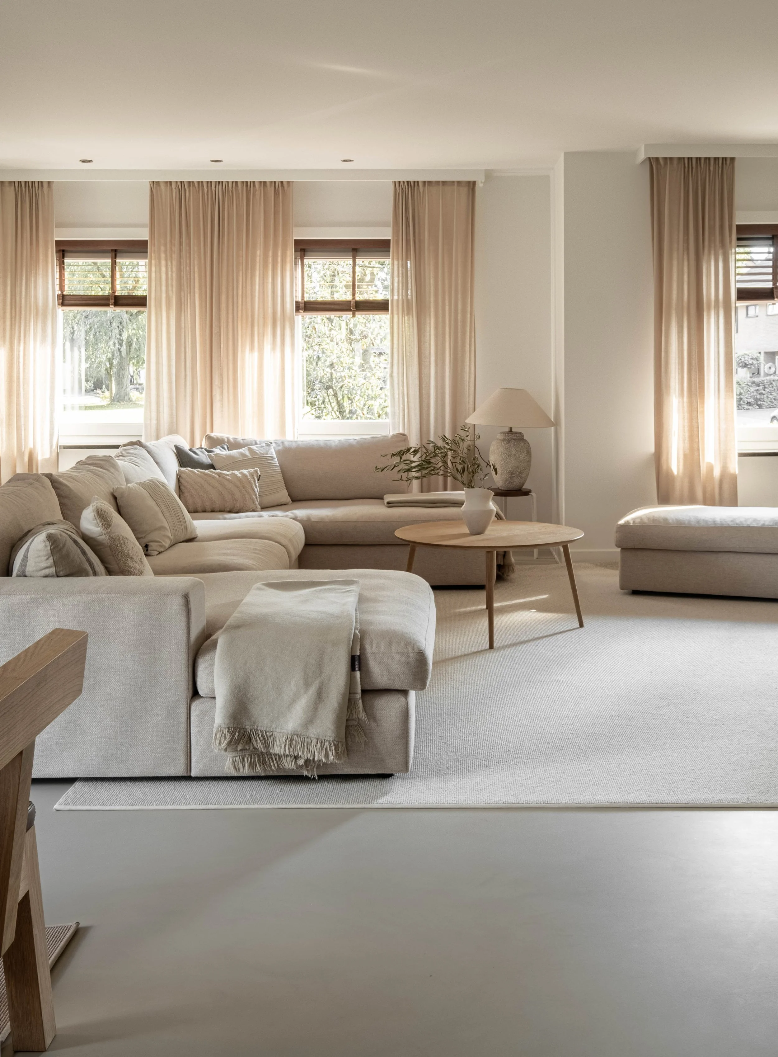 A cozy, well-lit living room with beige and cream-colored furniture, curtains, and decor, featuring a large sectional sofa, a round wooden coffee table, and large windows with beige curtains.