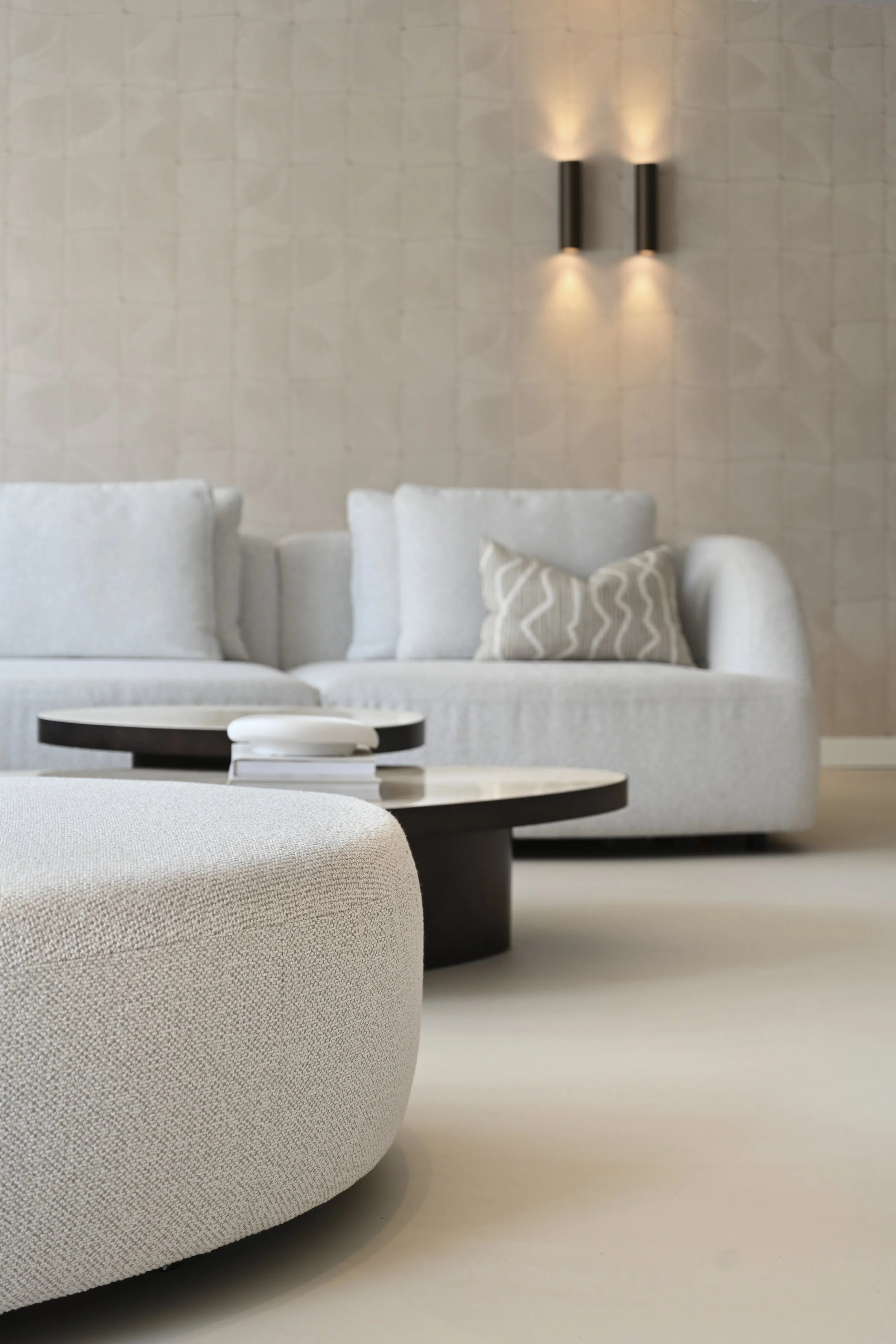 Modern living room with a light gray sofa, black and white coffee table, beige ottoman, and beige wall with two black wall sconces