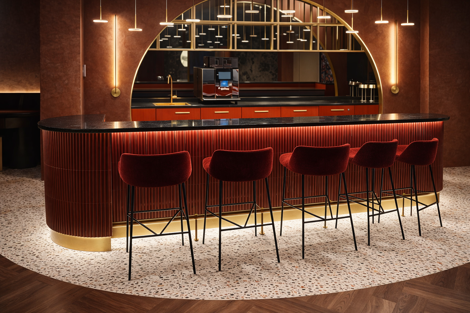 A stylish bar with a curved, ribbed red front, black marble countertop, and five red velvet bar stools with black metal legs. Behind the bar, there is a large mirror with a gold framing, reflecting the lighting fixtures and part of the kitchen area, which includes a modern espresso machine and gold accents. The space has warm, ambient lighting and a terrazzo floor with a curved wooden border.
