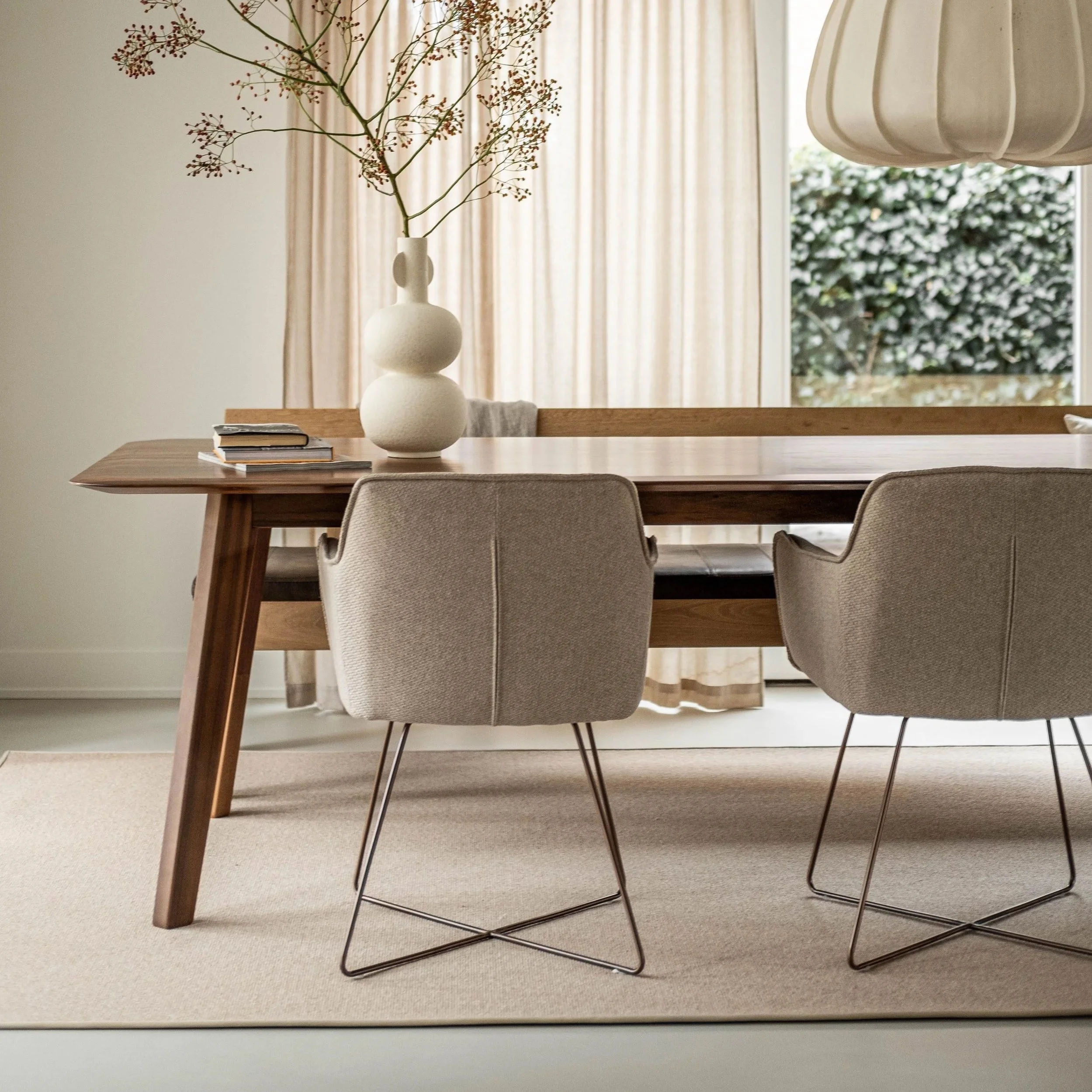 A modern dining room with a wooden table, two beige upholstered chairs with metal legs, a large white vase with a branch, and a window with beige curtains letting in natural light.