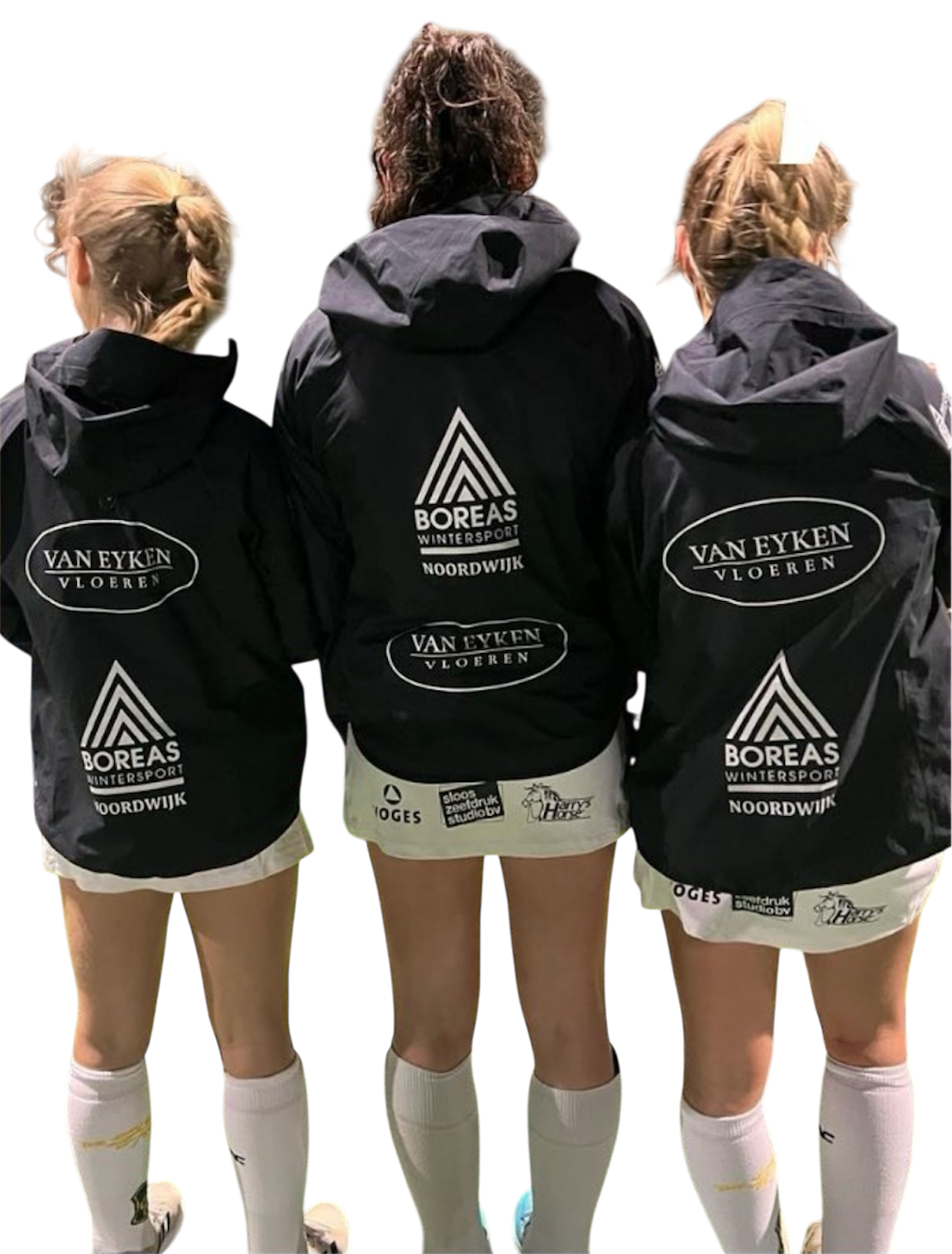 Three soccer players standing with their backs to the camera, wearing black jackets with logos and white shorts and socks.