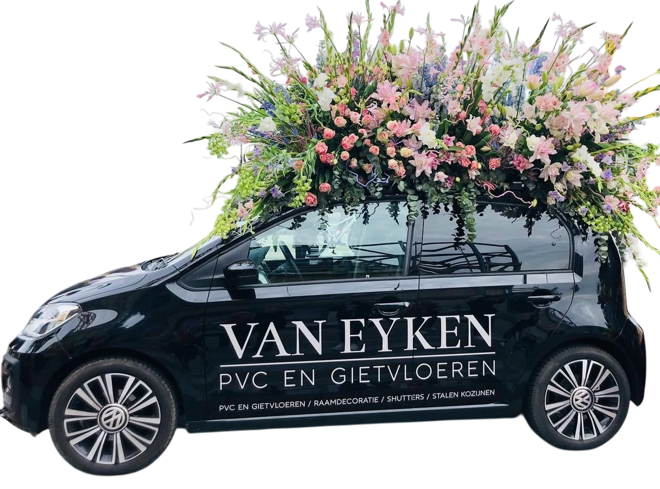 A small black Volkswagen car decorated with a large, colorful floral arrangement on top featuring pink, purple, and white flowers. The car has advertising text on the side for Van Eyken, a company specializing in PVC and ceramic flooring, window decoration, shutters, and steel frames.