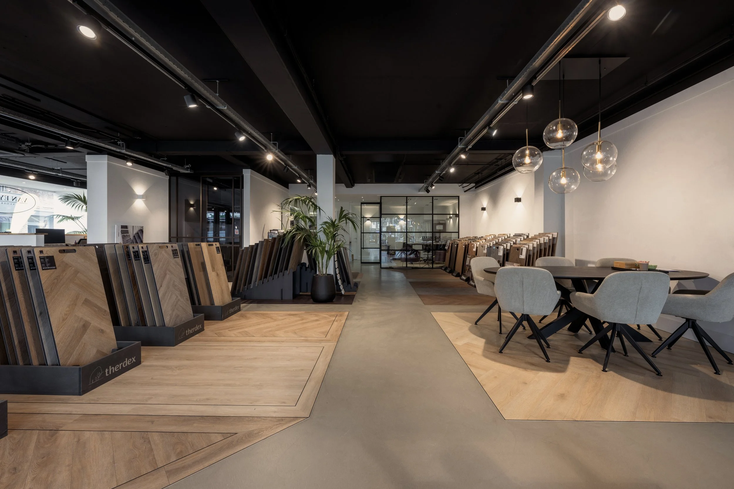 Interior view of a modern showroom with display of wooden flooring samples, seating area with beige chairs and table, and decorative lighting fixtures.