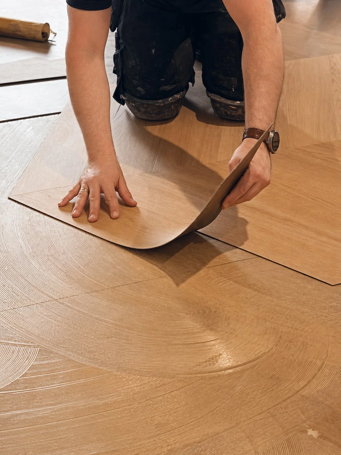 Een persoon legt een houten vloer met behulp van een vinyl ondergrond en een leggereedschap, gekleed in zwarte kleding met een horloge.