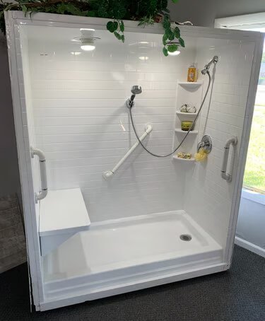 A white indoor shower stall with a built-in bench, handheld showerhead, and wall-mounted shelves holding toiletries, with a sliding glass door and a window outside.