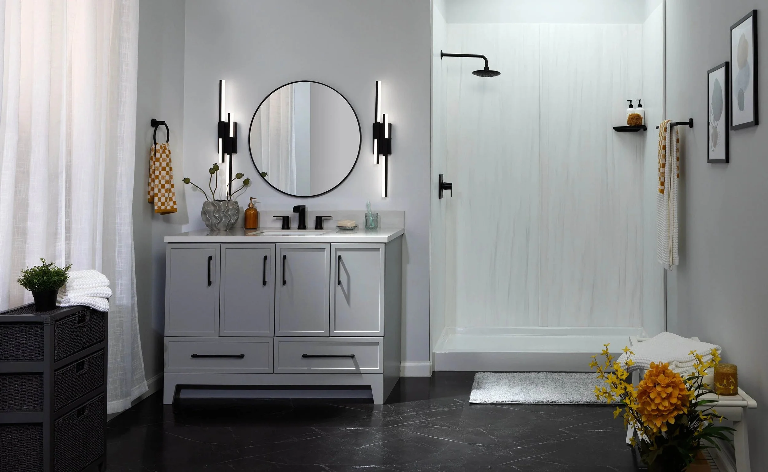 Modern bathroom with a white vanity, round mirror, black fixtures, and a walk-in shower with a black showerhead, black towel bar, and decorative framed art on the wall. The bathroom has white walls, black flooring, and yellow flowers for decoration.