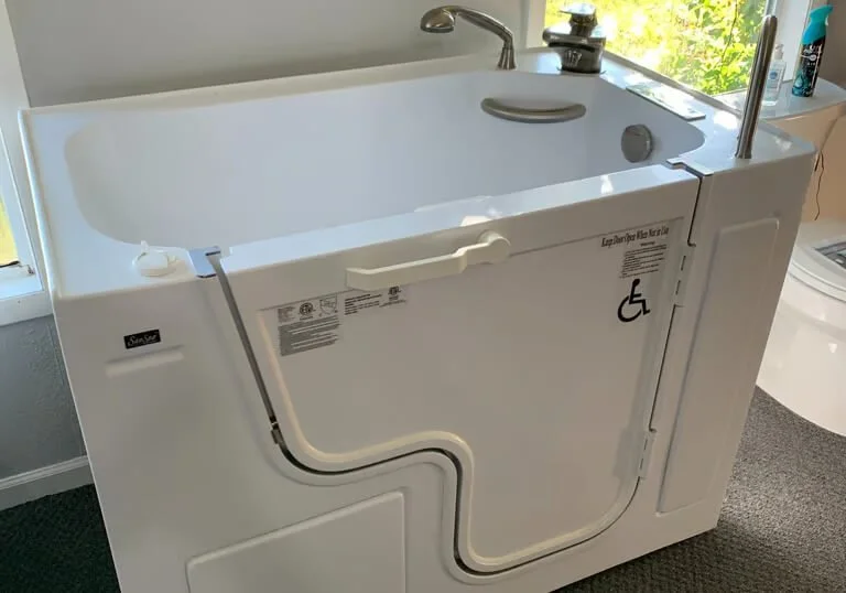 A medical walk-in bathtub with a door and a fold-down seat, designed for accessibility, located in a bathroom next to a window and laundry area.