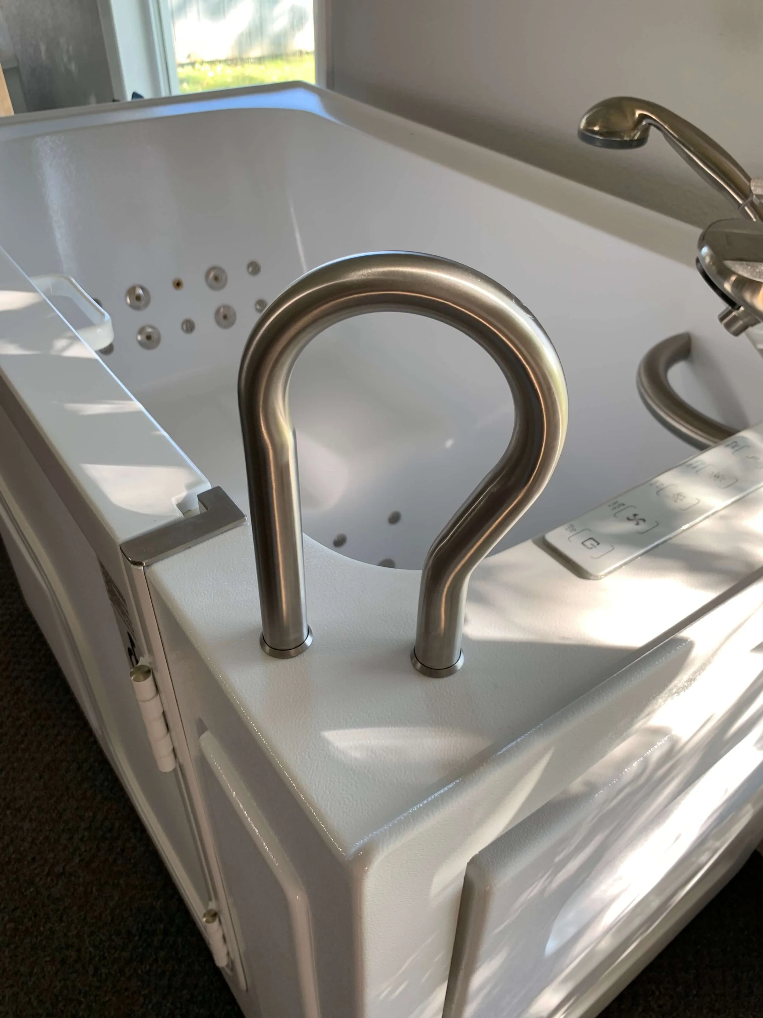 A white medical or therapeutic bathtub with stainless steel grab bars, multiple adjustable jets inside, and a control panel attached, placed near a window.