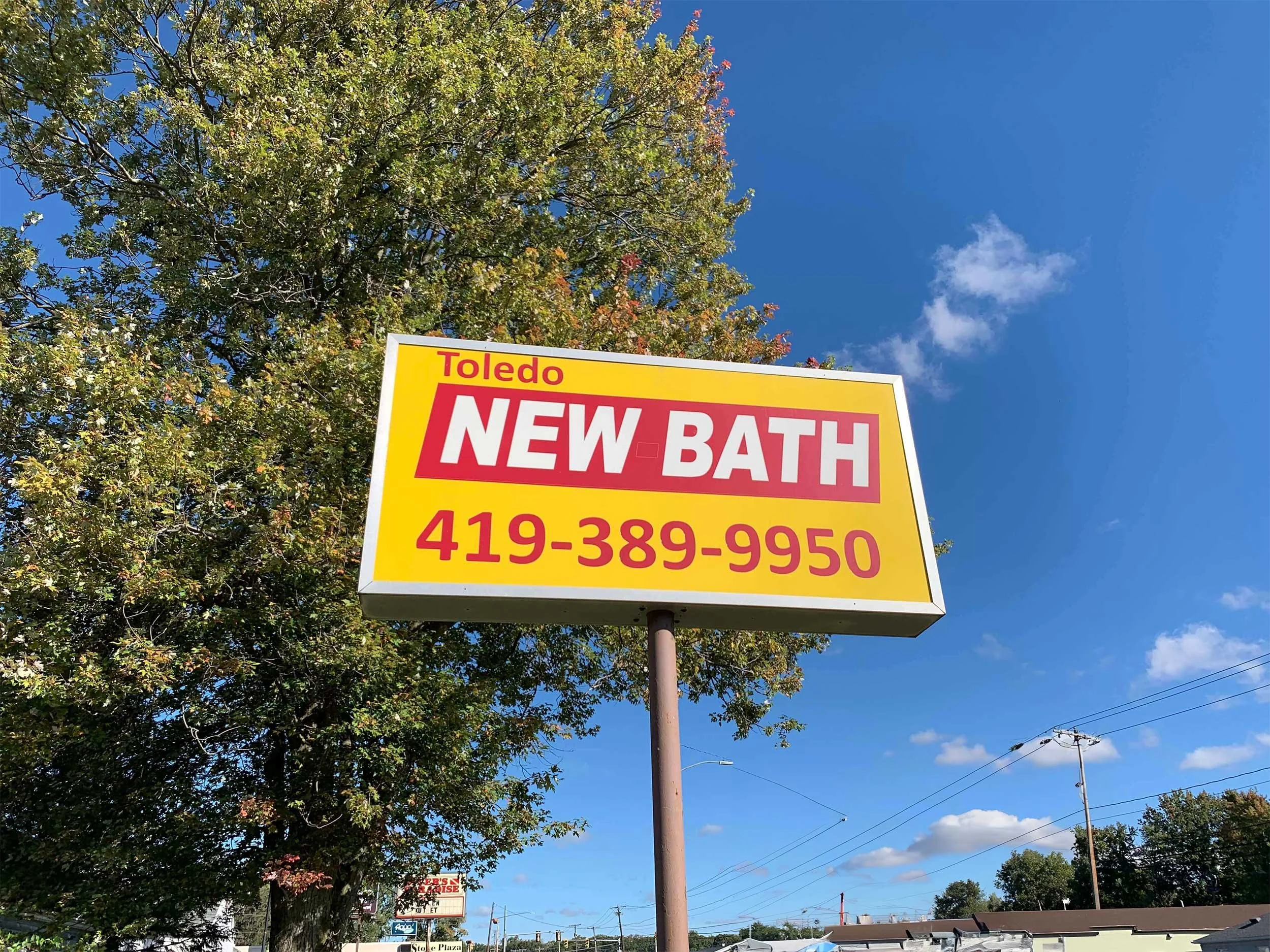 A bright yellow billboard with red and white text advertising a new bath service in Toledo, along with a phone number. It is set against a blue sky with some clouds and a tree nearby.