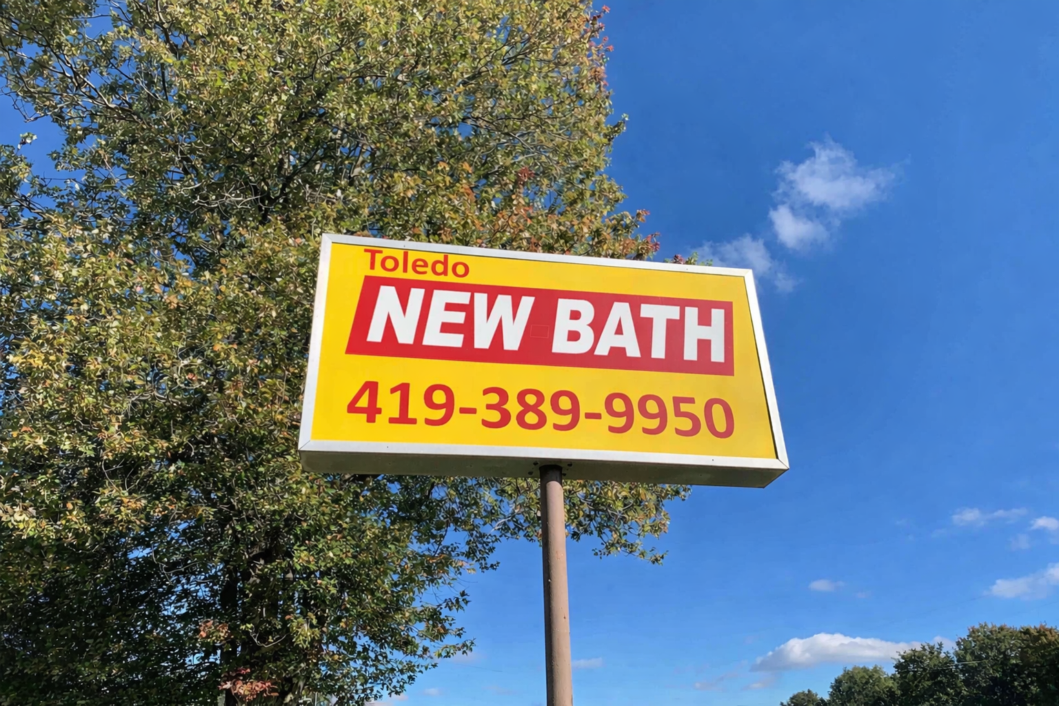 Sign for New Bath in Toledo with phone number 419-389-9950, against a background of trees and a blue sky with some clouds.