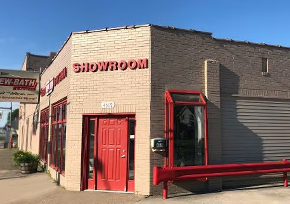 Building with a sign that reads 'SHOWROOM' in red letters, with a red door and large windows, and a gray roller shutter on the side. Part of a street with a sidewalk.