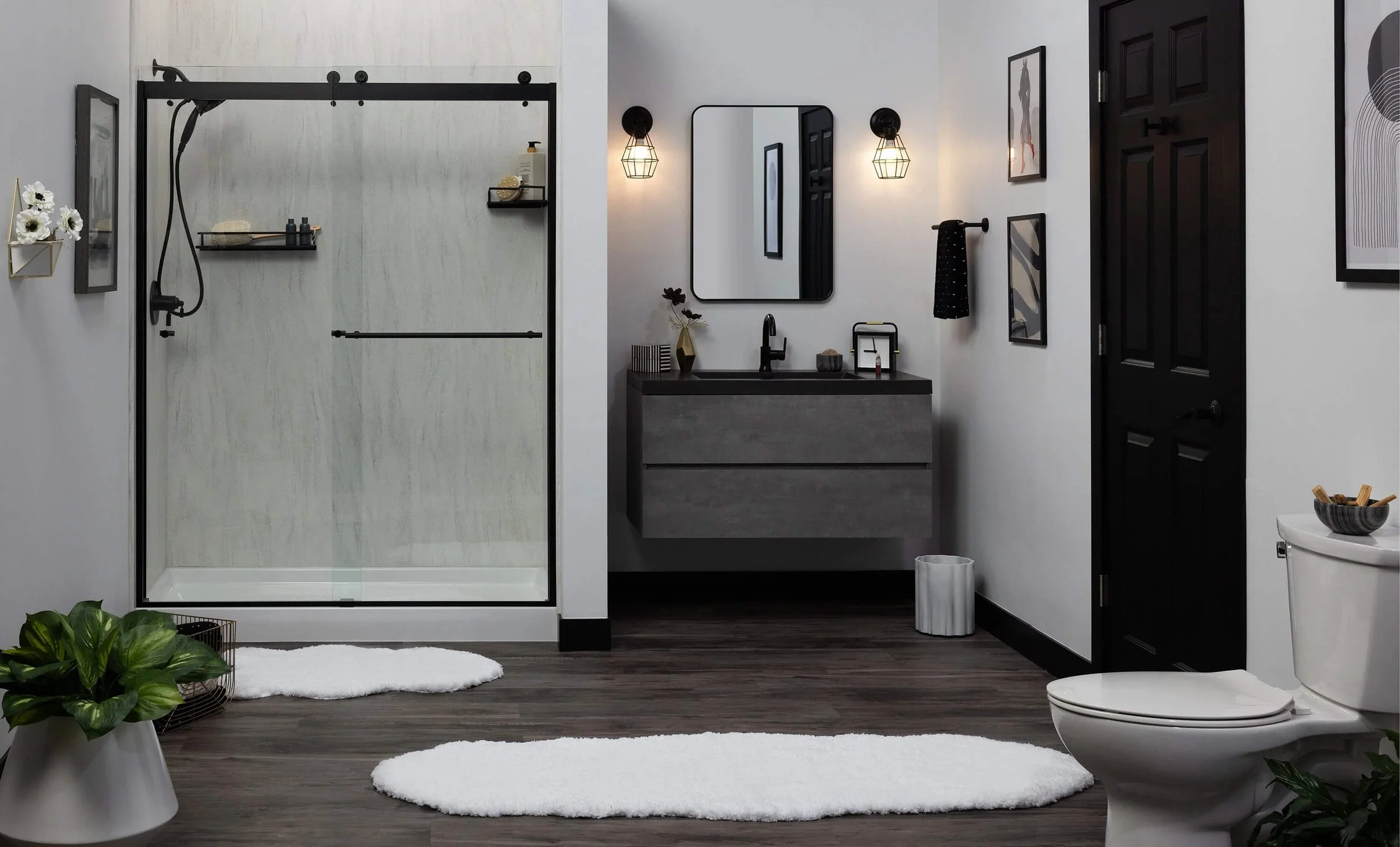Modern bathroom with a walk-in shower, gray vanity with a black faucet, wall mirror, black wall lights, framed art, black door, toilet, plants, and white rugs on dark wood floor.