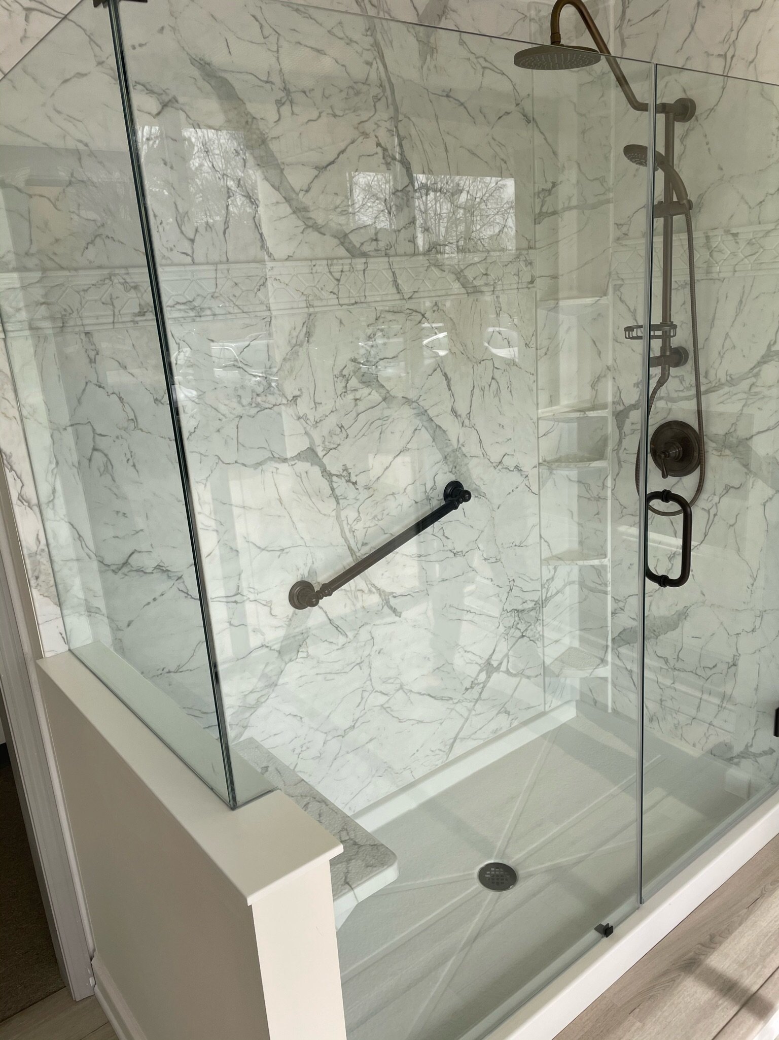 A walk-in shower with two showerheads, a glass door, marble walls, a built-in shelf, and a grab bar.