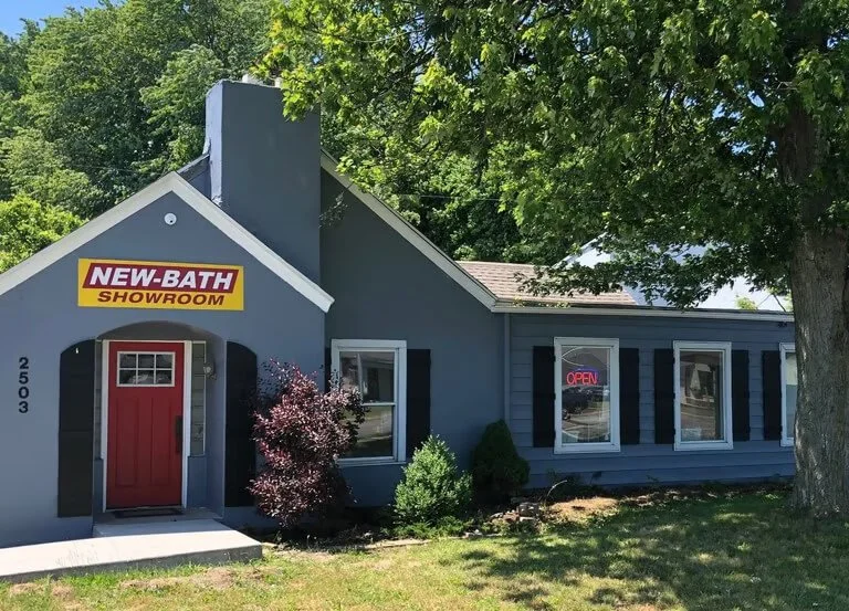 Blue house with a red door, a large tree on the right, and a sign that reads 'New-Bath Showroom' above the door, with a smaller 'Open' sign in the window.
