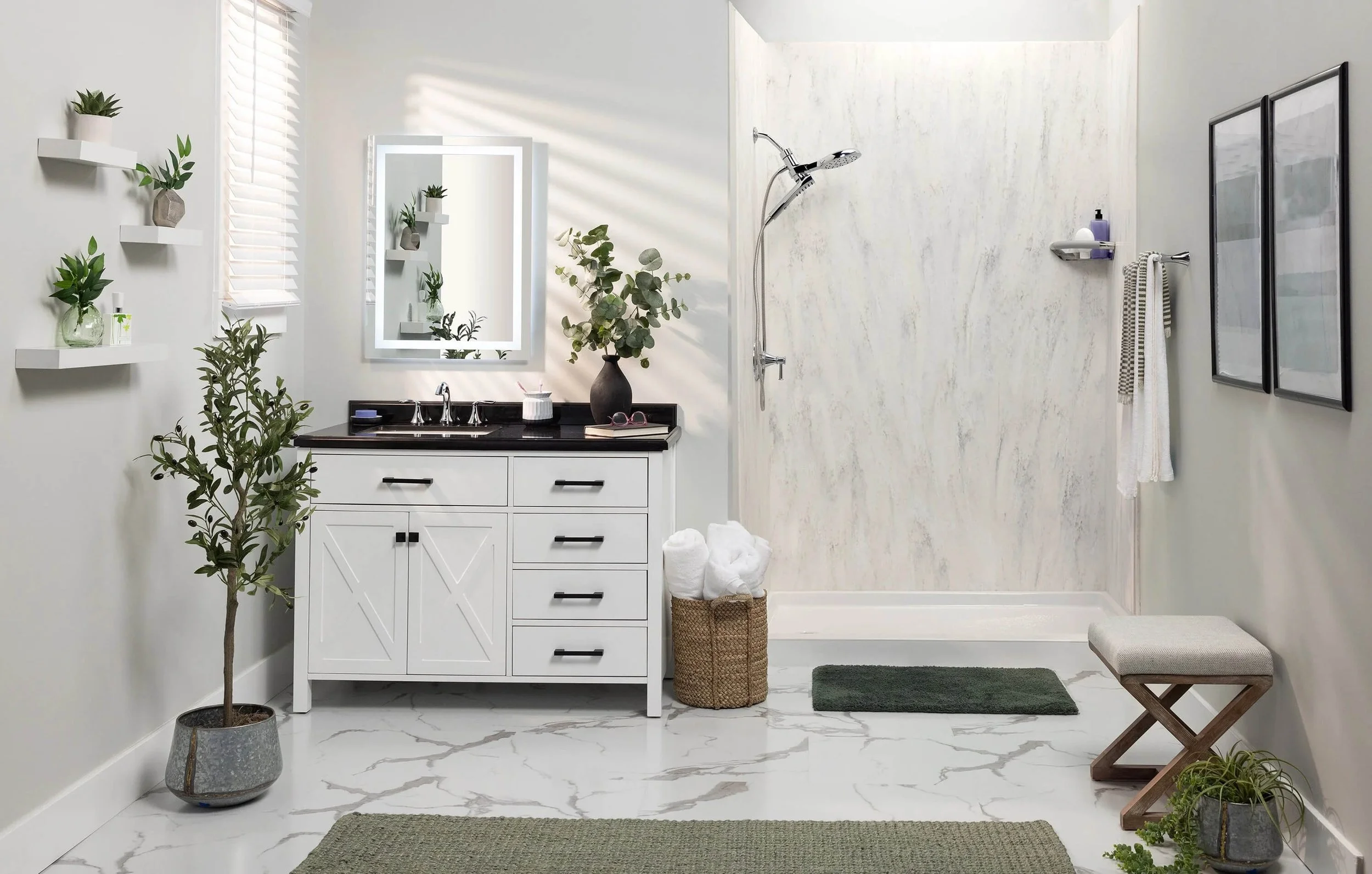 A modern bathroom with a white vanity with black countertop, potted plants, a mirror with backlighting, and a walk-in shower with a textured wall. Decor includes framed art, a small stool, and hanging shelves with plants.