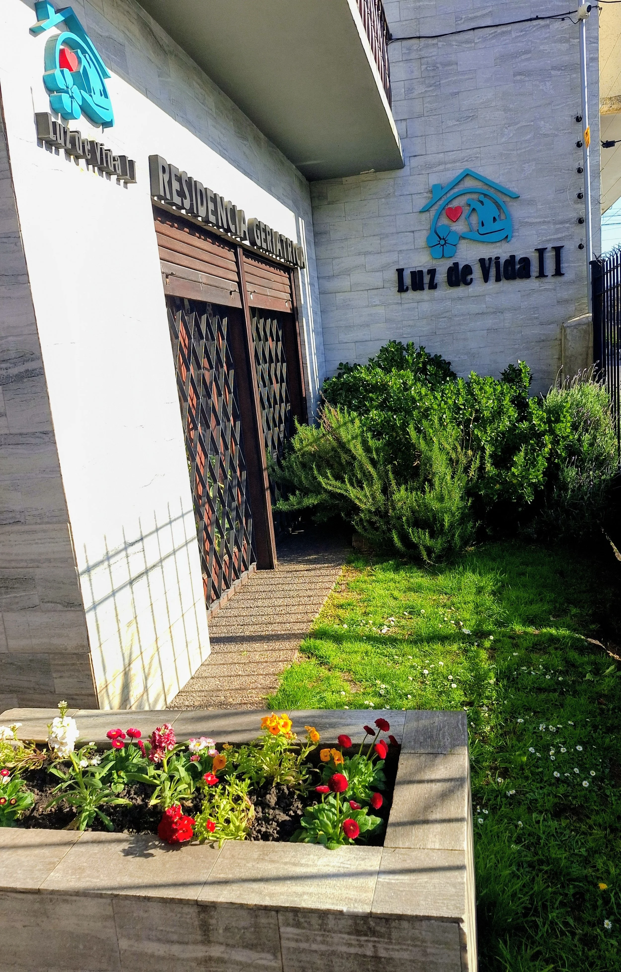 Entrada a un edificio llamado Luz de Vida II con plantas y flores en un macetero, y un muro de ladrillos con un logo que combina una casa, un corazón, una flor y una ola.
