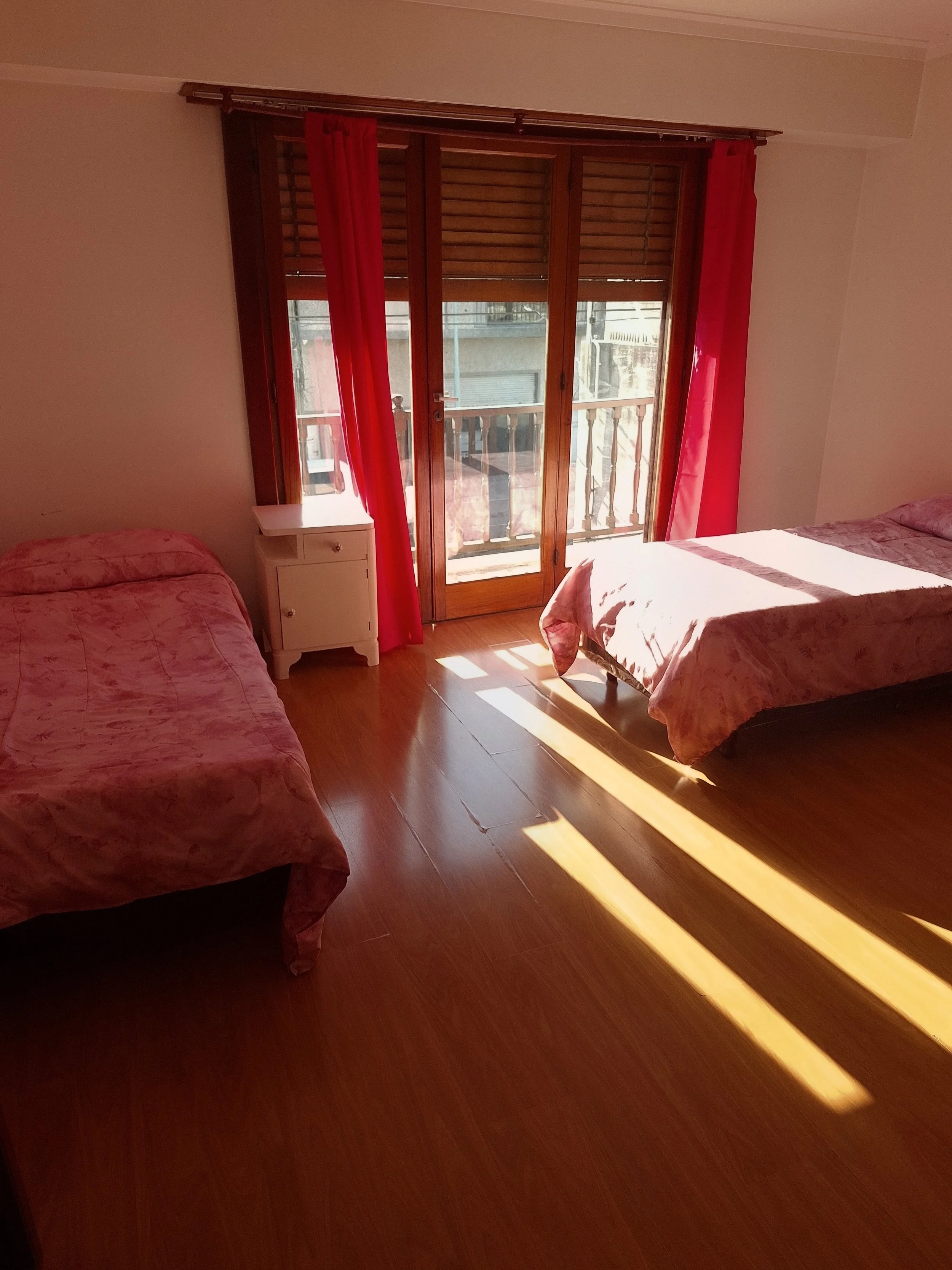 Habitación con dos camas con ropa de cama rosa, ventanas grandes con cortinas rojas y persianas de madera, y una pequeña mesa de noche blanca.