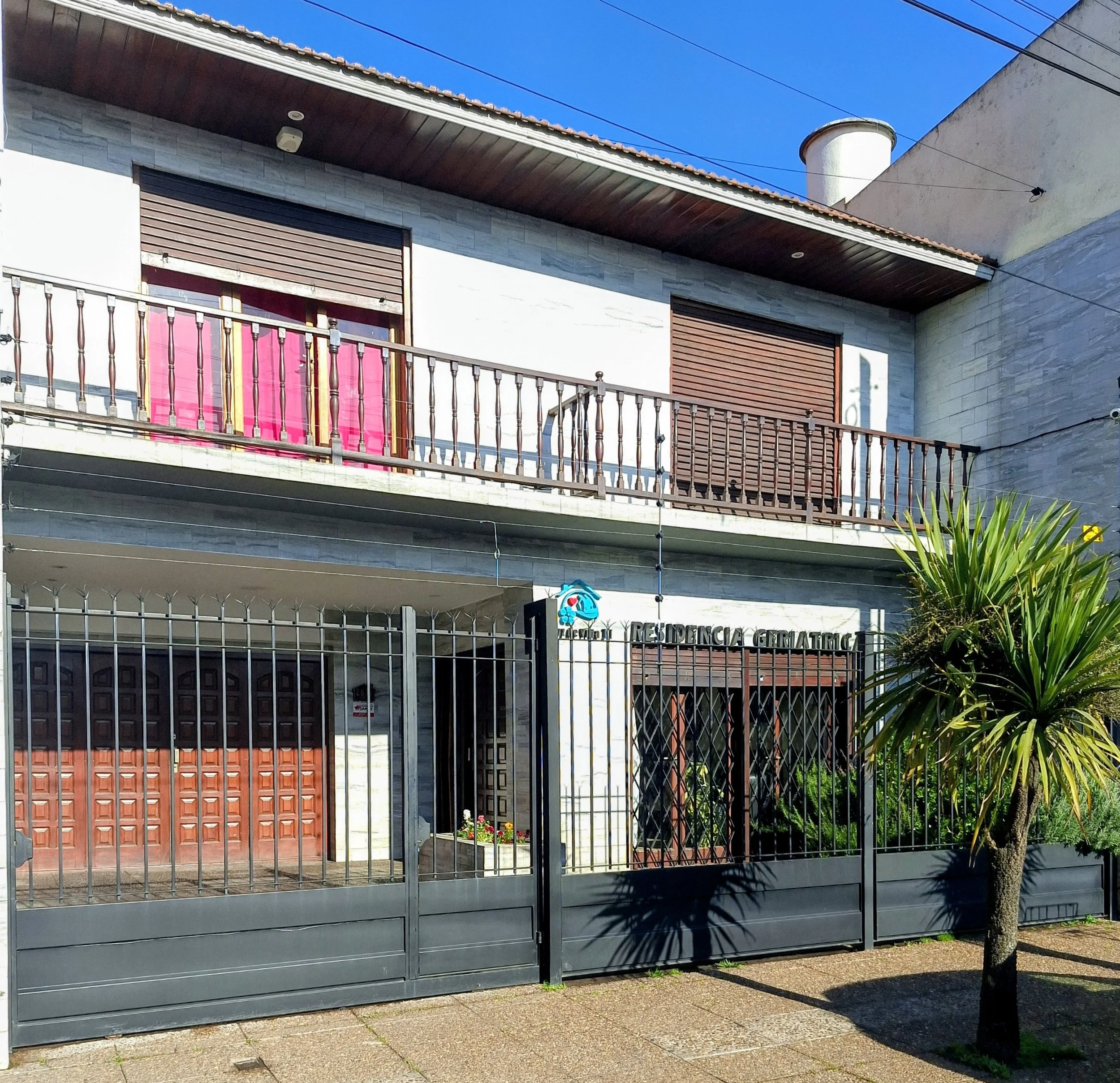 Vista de un edificio residencial con rejas en la entrada, balcón con cortinas rosas y paredes de piedra, en un día soleado con cielo despejado azul.