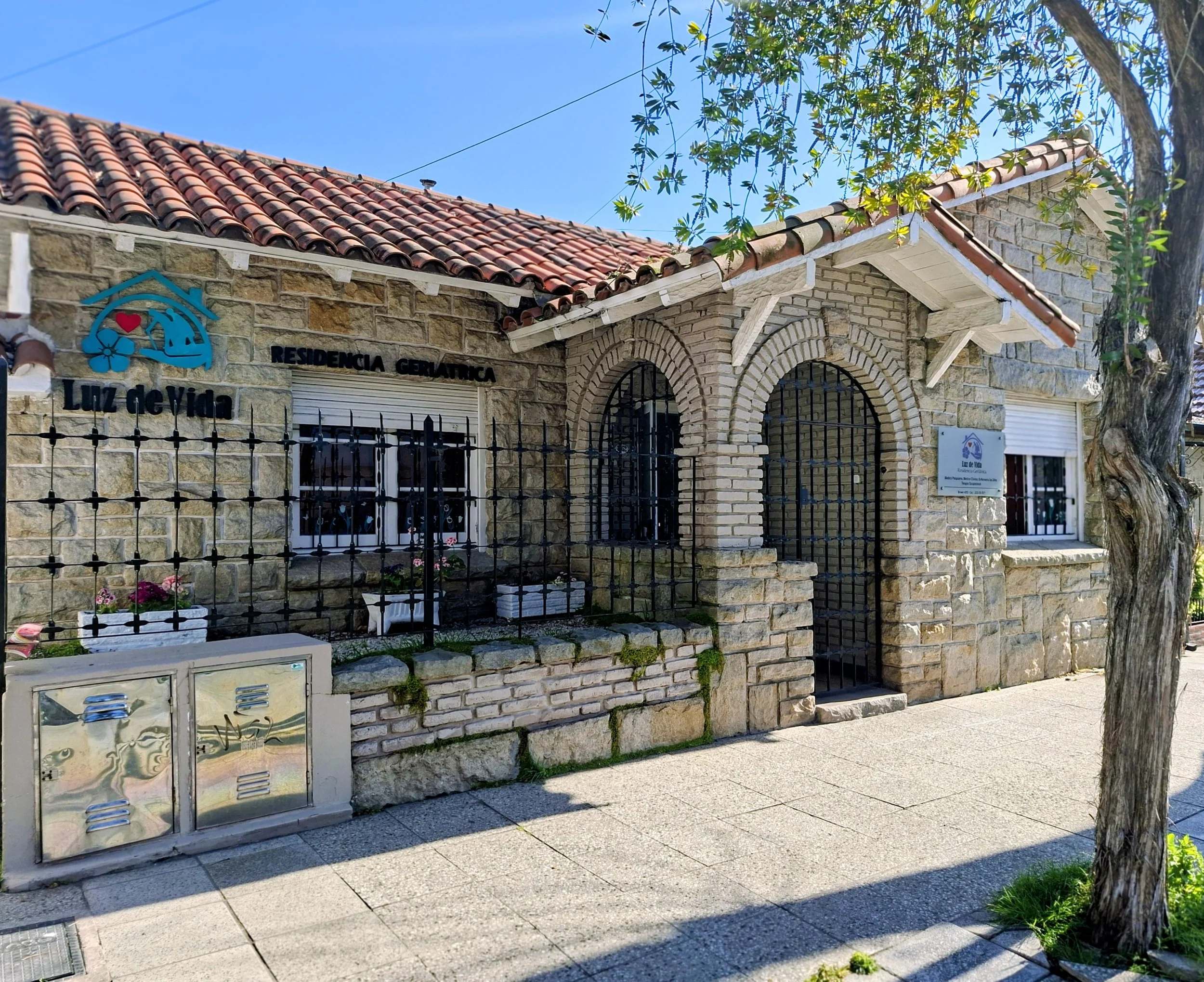 Fachada de una residencia con paredes de piedra y ladrillo, techo de tejas rojas, rejas negras en las ventanas, y cartel que dice 'Residencia Gerlática Luz de Vida'.