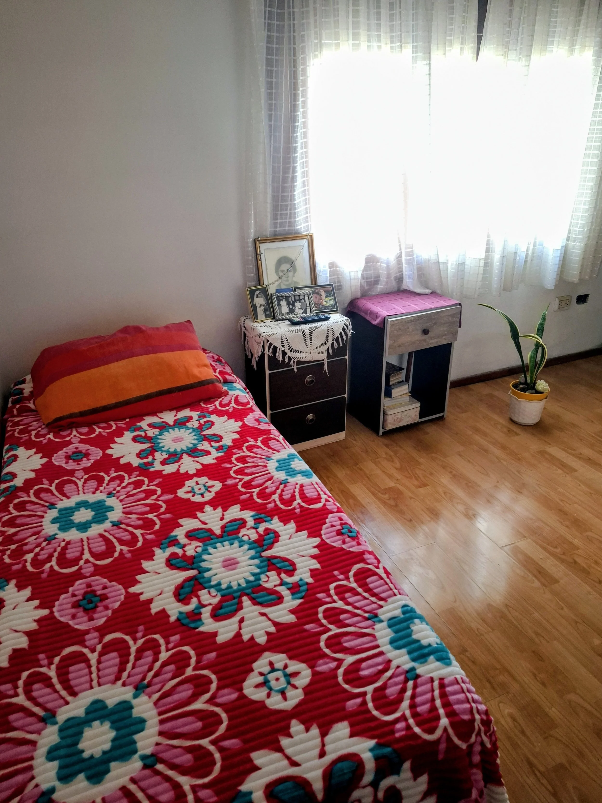 Una habitación con una cama con cobertor rosa con patrones de flores y un almohadón naranja. Hay una pequeña mesa con fotos, control remoto y un cuadro. También hay un mueble con libros y un florero con planta, y una ventana con cortinas blancas y de cuadros. La habitación tiene piso de madera.