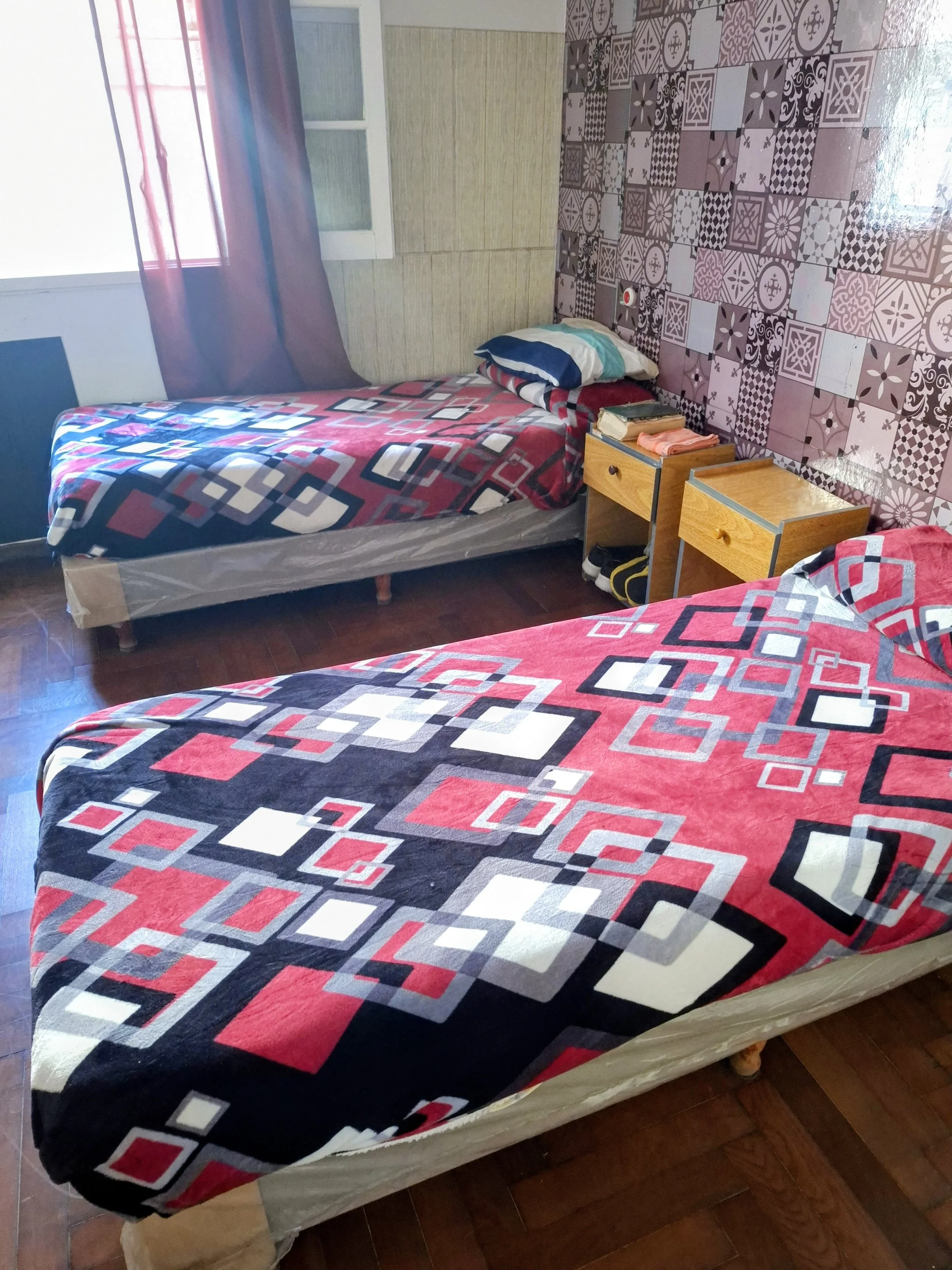 La imagen muestra dos camas con colchas de colores rojo, negro, gris y blanco con patrones geométricos. Entre las camas hay una mesita de madera con dos cajones, en la que hay varios objetos, incluyendo un libro, un par de toallas pequeñas y una bolsa. La pared de fondo tiene un diseño de baldosas en tonos morados, rosas, grises y blancos con diferentes patrones. Hay una ventana con cortinas moradas que deja pasar la luz hacia la habitación. La habitación tiene suelo de madera.