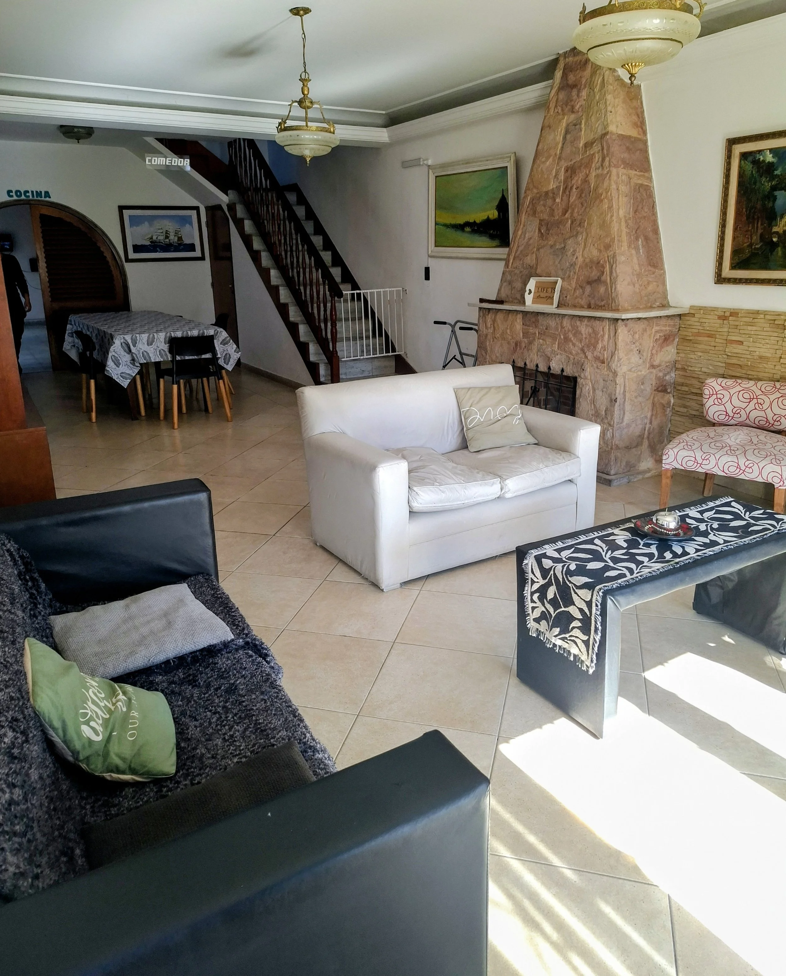 Sala de estar con sillones y una chimenea de piedra, cuadro en la pared y una mesa de centro con un adorno. Escalera de madera lleva a otro piso.