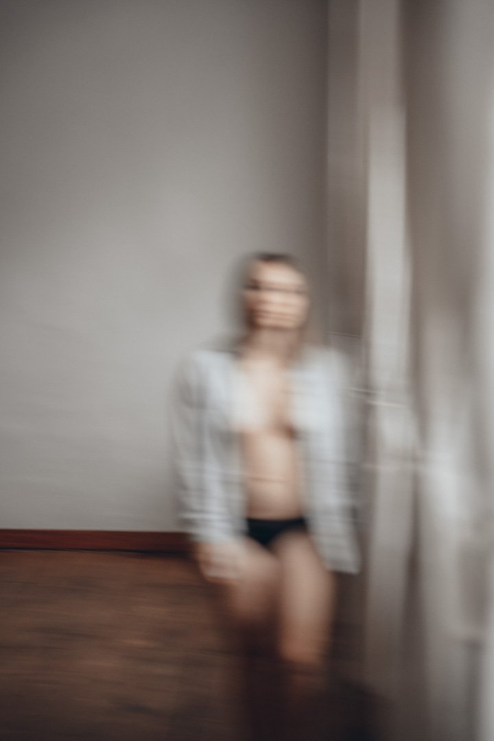 Ein verschwommenes Bild einer Frau, die vor einem neutral-coloreden Raum steht, teilweise hinter einem Vorhang verborgen, trägt dunkle Unterwäsche und eine weiße Jacke.