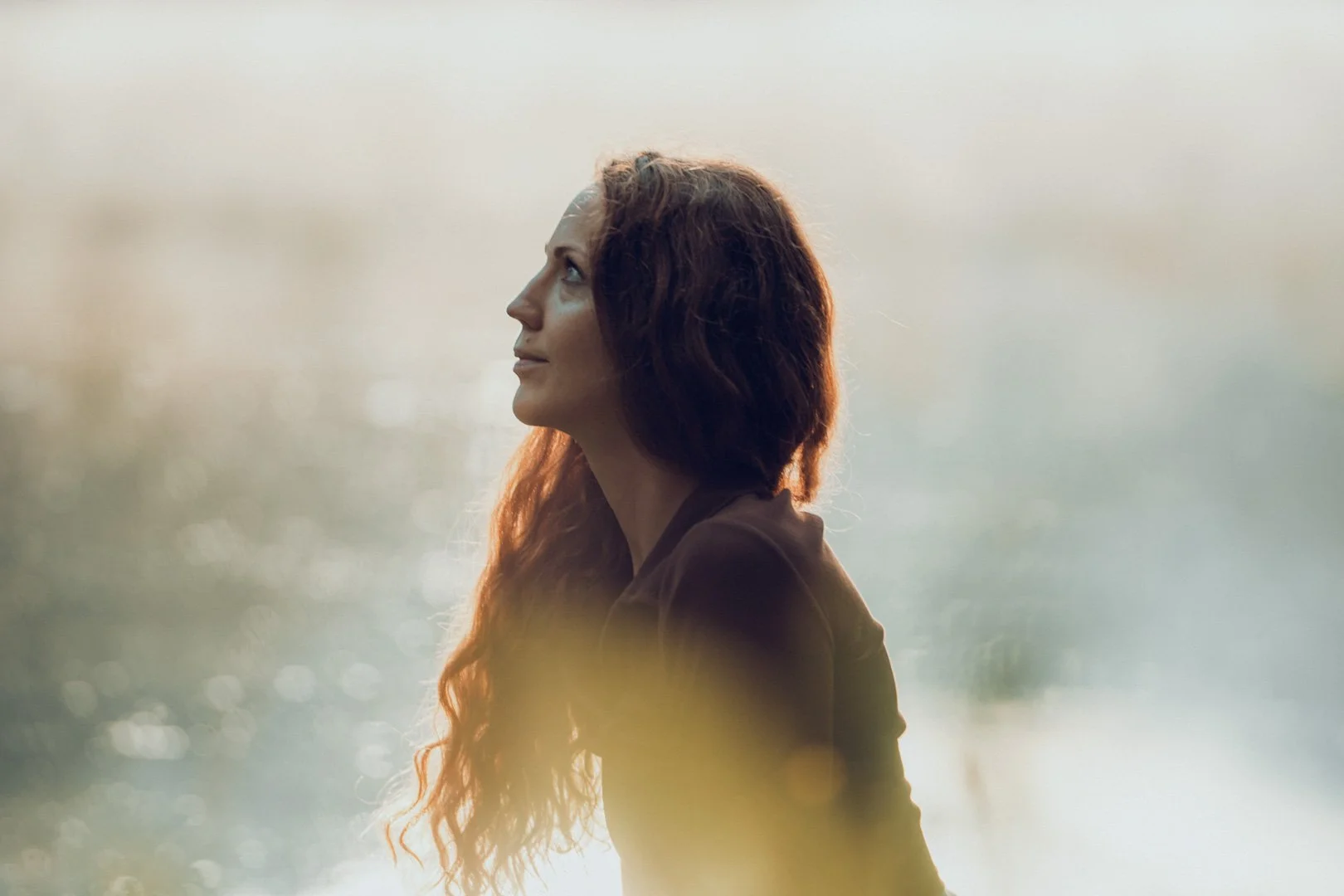 Eine Frau mit lockigem, braunem Haar, die nach oben schaut, vor einem unscharfen natürlichen Hintergrund.