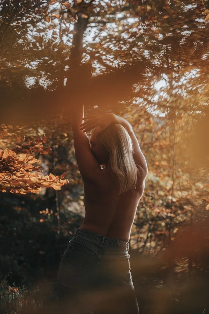 Ein nackt, blonde Frau steht in einem Herbstwald mit goldbraunen Blättern, hebt die Arme und positioniert die Hände vor dem Gesicht.