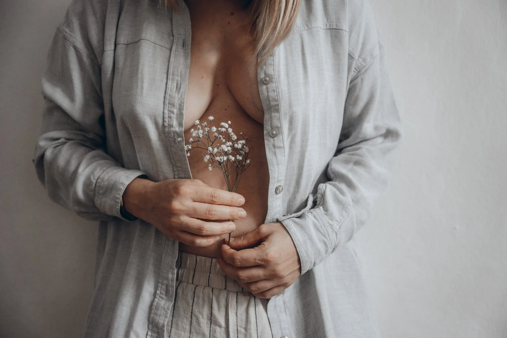 Nahaufnahme eines Frauenoberkörpers, die Frau trägt ein offenes, helles Hemd und hält eine kleine Blumenstrauß aus weißen Blumen an der Brust.