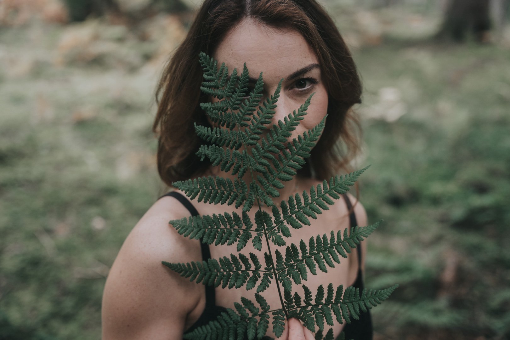Eine Frau steht im Freien, hält ein grünes Farnblatt vor ihr Gesicht und schaut durch das Blatt.