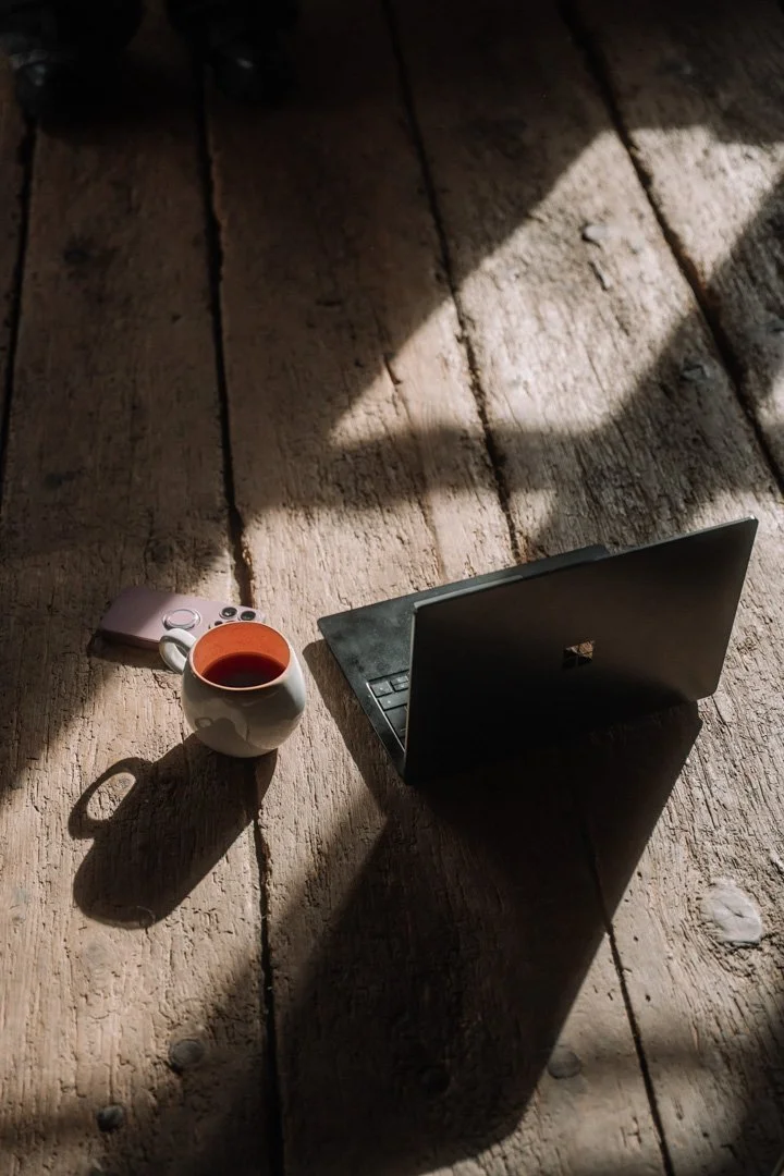 Ein offenes Laptop, eine Tasse Tee, eine Handy und eine Fernbedienung auf einem Holztisch im Sonnenlicht.