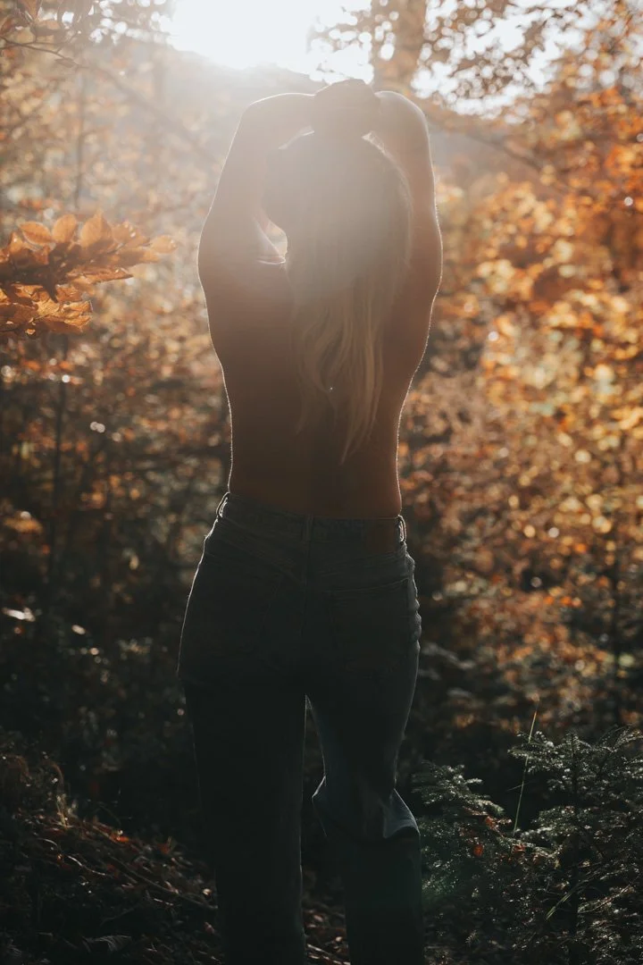 Eine nackte Frau steht in einem Wald bei Sonnenaufgang oder Sonnenuntergang, mit Sonnenlicht im Hintergrund und herbstlichen Blättern.