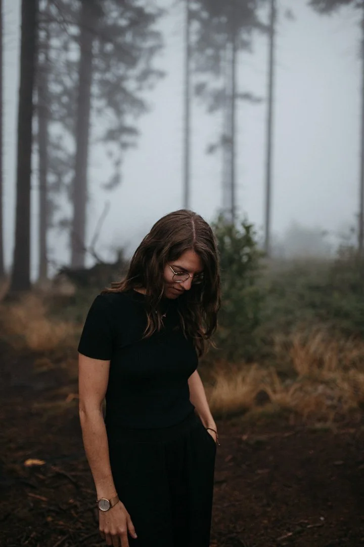 Junge Frau mit Brille und dunklem Outfit steht in einem nebligen Wald, schaut nach unten.