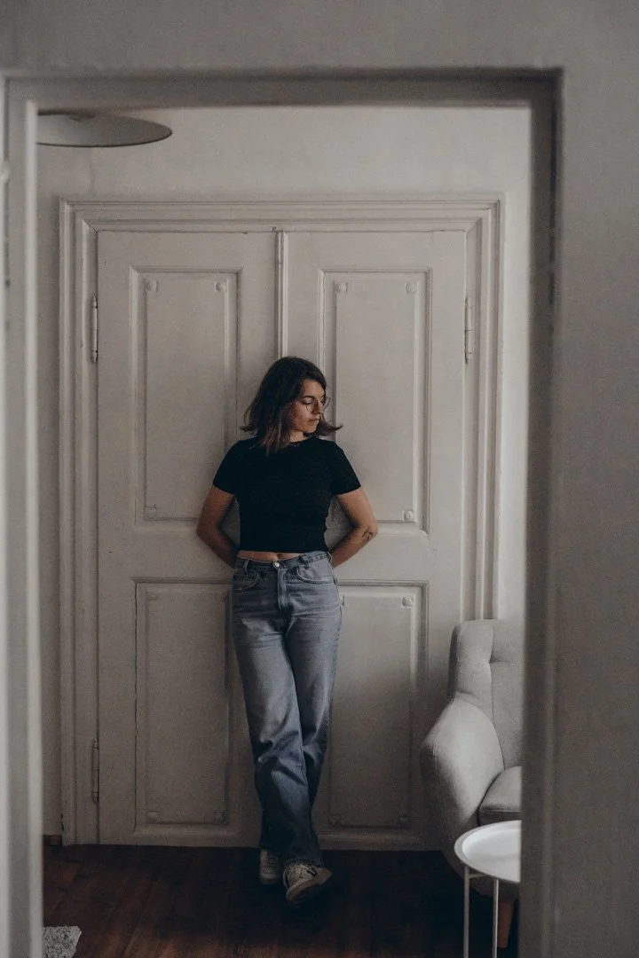 Junge Frau steht vor einer weißen Tür, trägt ein schwarzes T-Shirt, Jeans und Brille, während sie in einem modernen Raum steht.