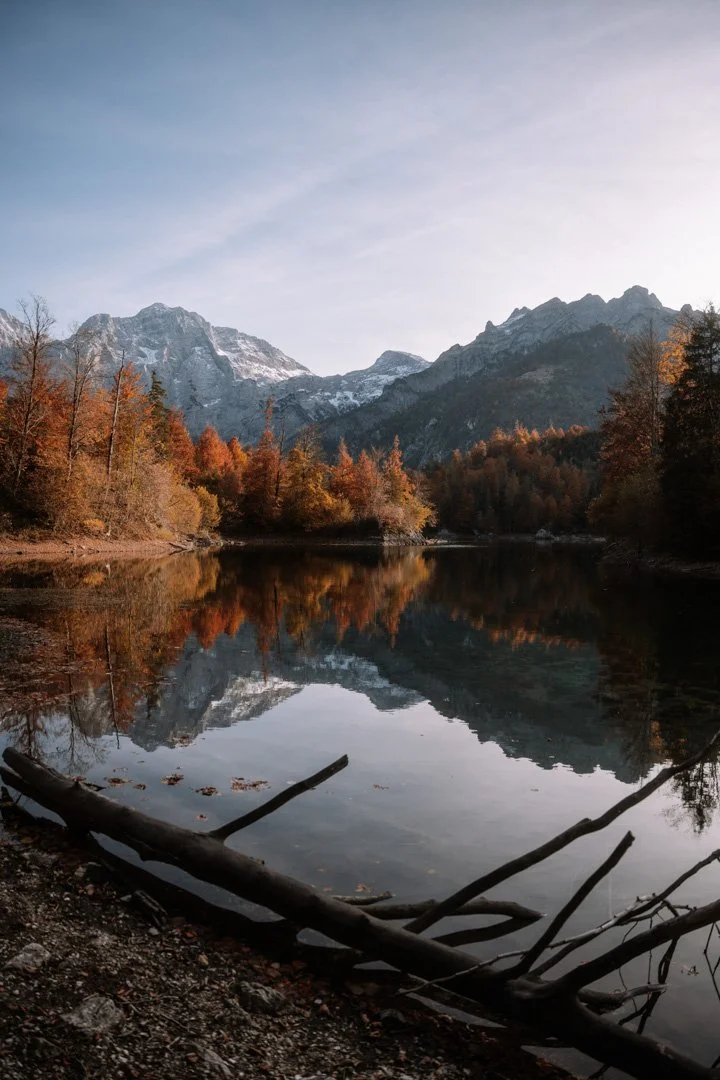 Ein ruhiger See spiegelt die umliegenden Berge und bunten Herbstbäume wider, im Vordergrund liegen einige Äste am Ufer.