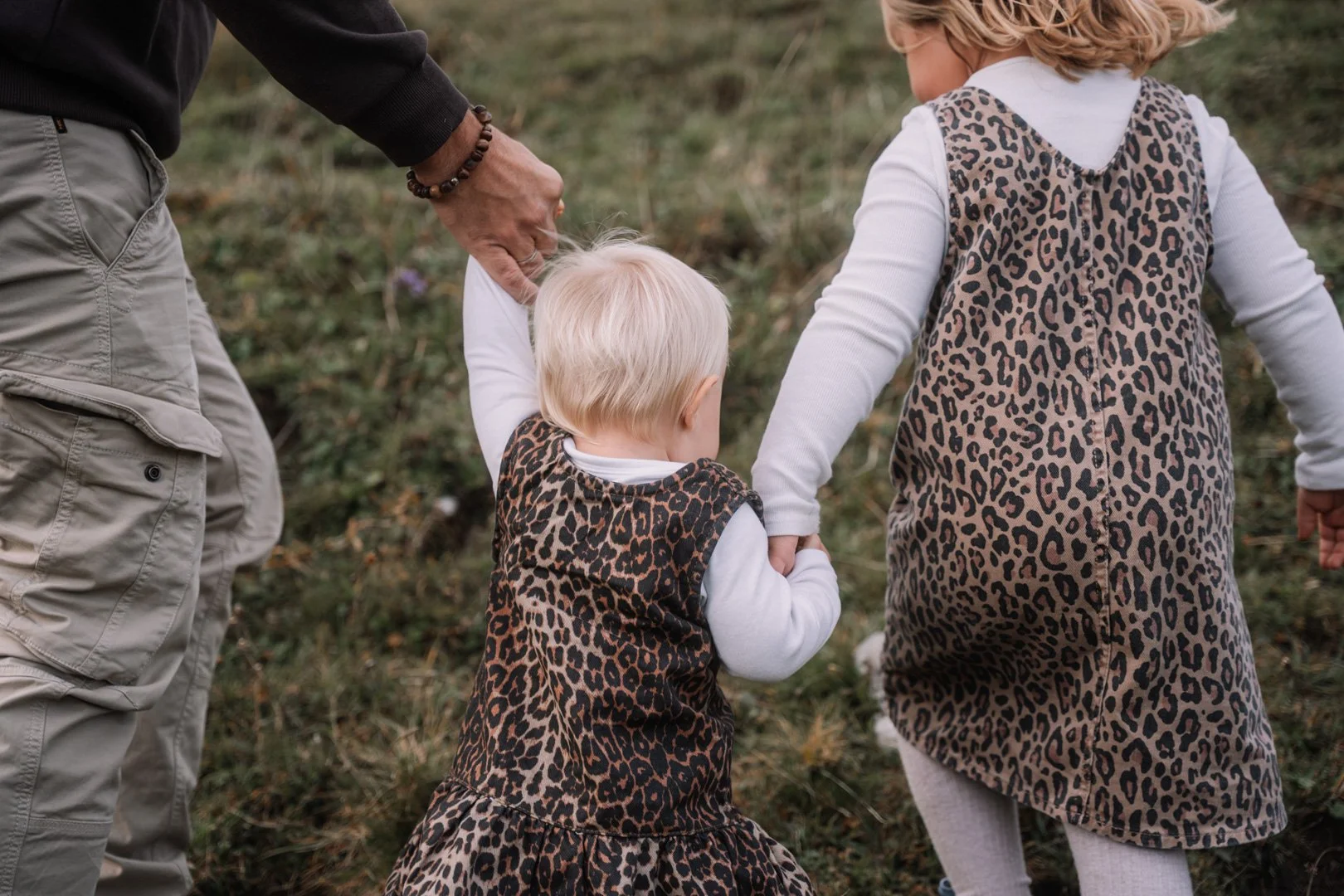Zwei Kinder, ein Mädchen und ein Junge, laufen mit einem Erwachsenen im Freien auf einem grasbewachsenen Platz, alle tragen Tierprint-Kleidung.
