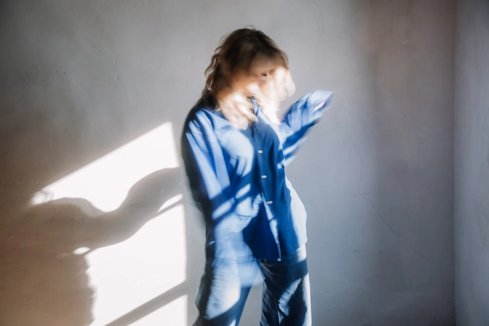 Eine junge Frau in blauer Hemd und Jeans steht gegen eine graue Wand, durch die Sonnenlicht und Schatten fallen.