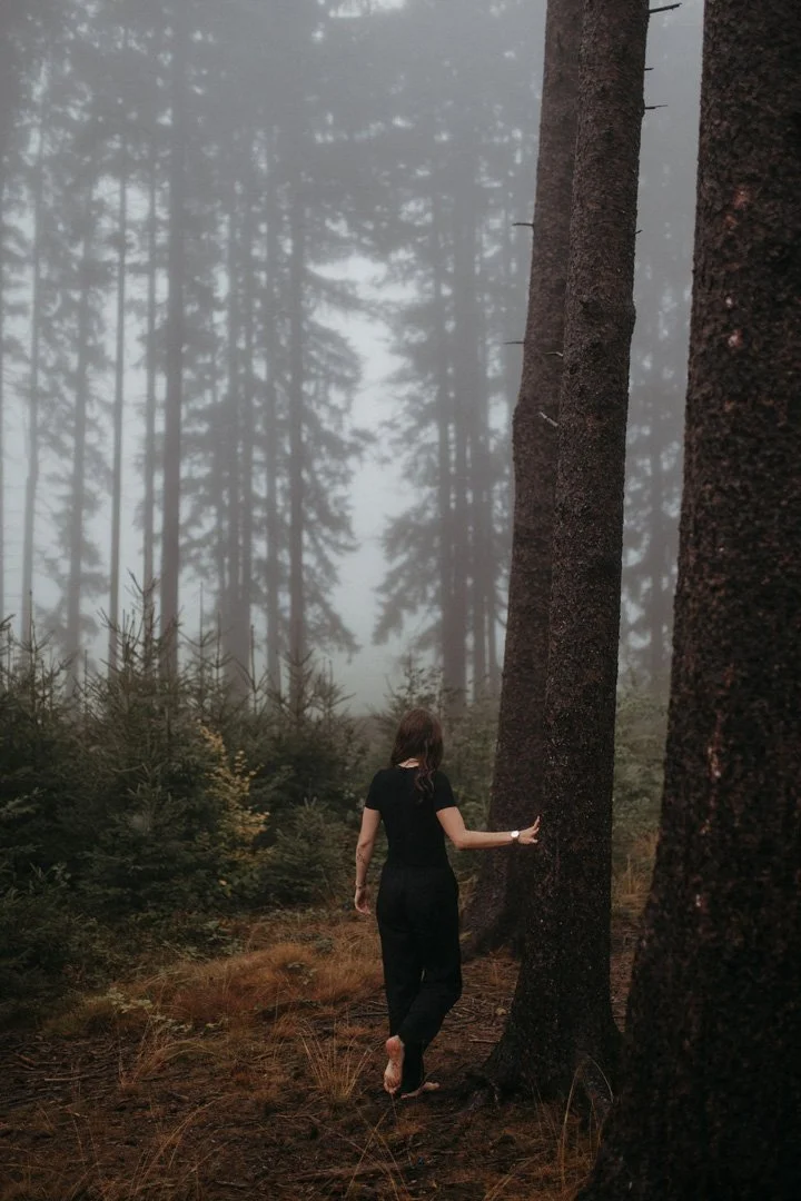 Eine Frau läuft barfuß durch einen nebelverhangenen Wald, umgeben von hohen Bäumen und dichtem Unterholz.