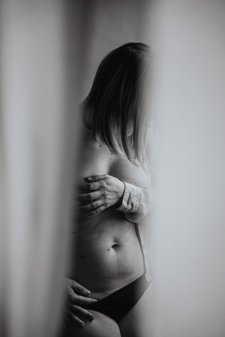 Schwarz-Weiß-Fotografie einer jungen Frau, die halb im Bild versteckt hinter einer Wand steht, mit einem Arm über die Brust und einem weiteren an der Taille, trägt dunkle Unterwäsche und hat mittellanges, gestreiftes Haar.