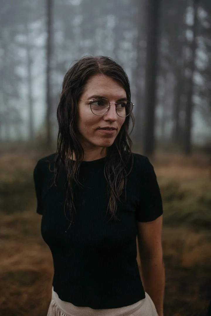 Junge Frau mit langen, nassen Haaren und Brille steht in einem nebligen Wald und schaut nach rechts.
