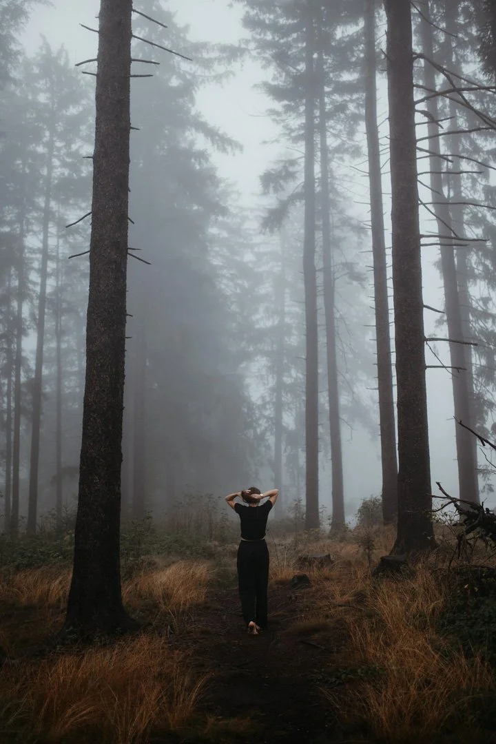 Eine Person in schwarzer Kleidung steht auf einem Waldweg, umgeben von hohen, dünnen Bäumen, im Nebel. Sie hat die Hände hinter dem Kopf.