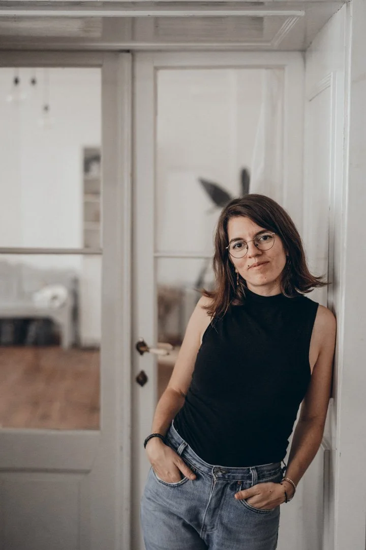 Junge Frau mit Brille, schwarzen Oberteil und Jeans lehnt an einer weißen Wand in einem hellen Raum mit Glas-Tür im Hintergrund.