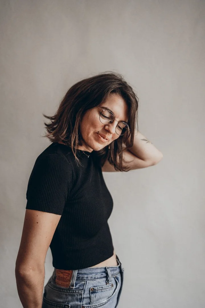 Eine junge Frau mit dunklem, mittellangem Haar, Brille, trägt ein schwarzes T-Shirt und Jeans, steht vor einer neutralen Wand, lächelt leicht, sieht nach unten.
