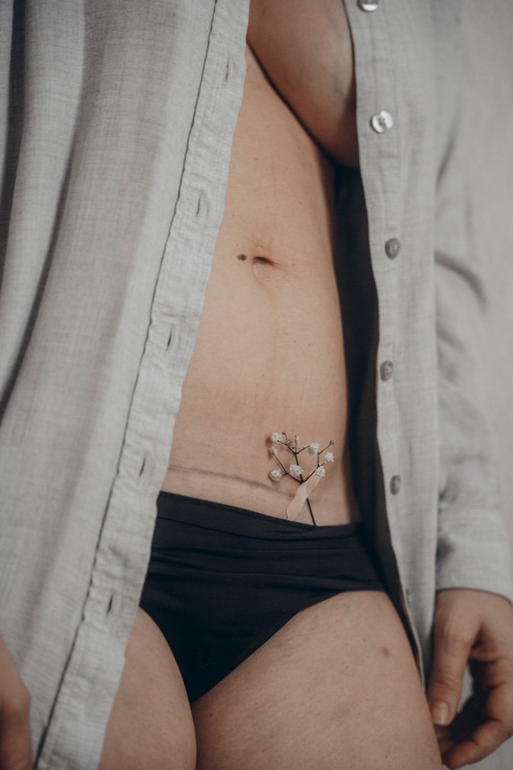 Nahaufnahme eines Torsos mit offener Hemdknopfleiste, schwarzer Unterwäsche und sichtbarem Bauchnabel, auf dem ein kleiner Tattoo-Schmuck mit Blumenmotiv befindet.