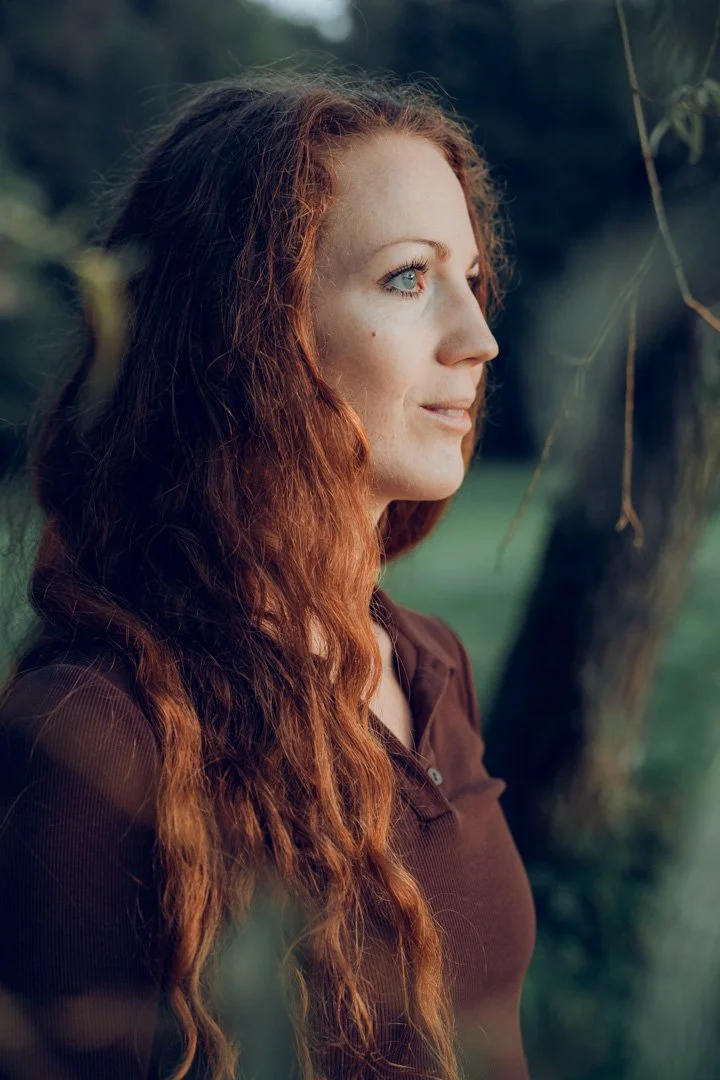 Nahaufnahme einer rothaarigen Frau im Profil vor einem Baum im Freien.