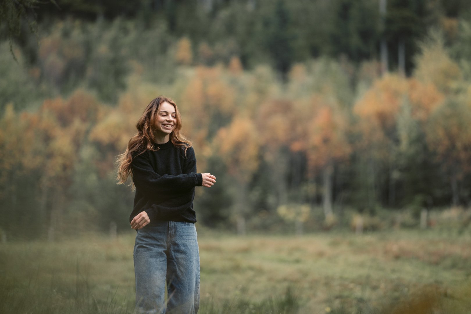 Junge Frau mit langen, lockigen Haaren, lachend im Grünen, im Herbst mit buntem Laub im Hintergrund.