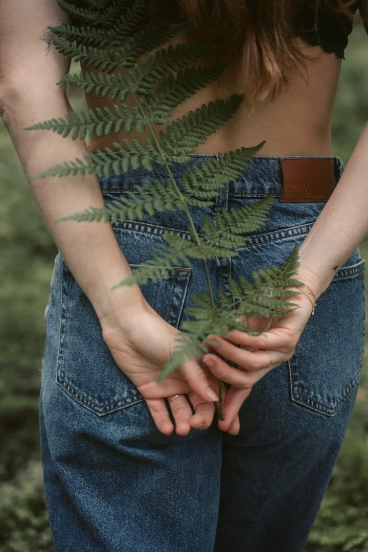 Eine Person in Jeans hält eine große grüne Farnpflanze hinter ihrem Rücken, im Hintergrund ist eine Naturkulisse zu sehen.