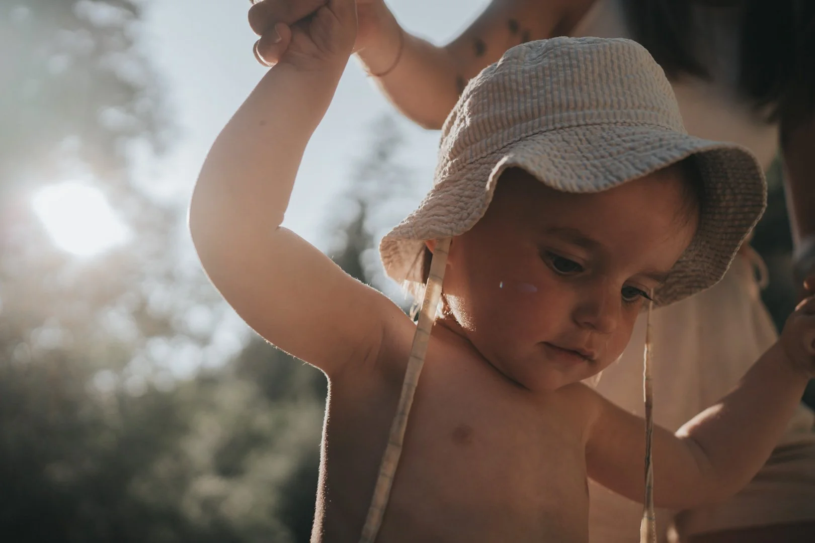Ein junger, nackter Junge trägt einen Sonnenhut und wird von einer Erwachsenenfigur gestützt. Es scheint warm und sonnig zu sein, mit Sonnenlicht im Hintergrund.