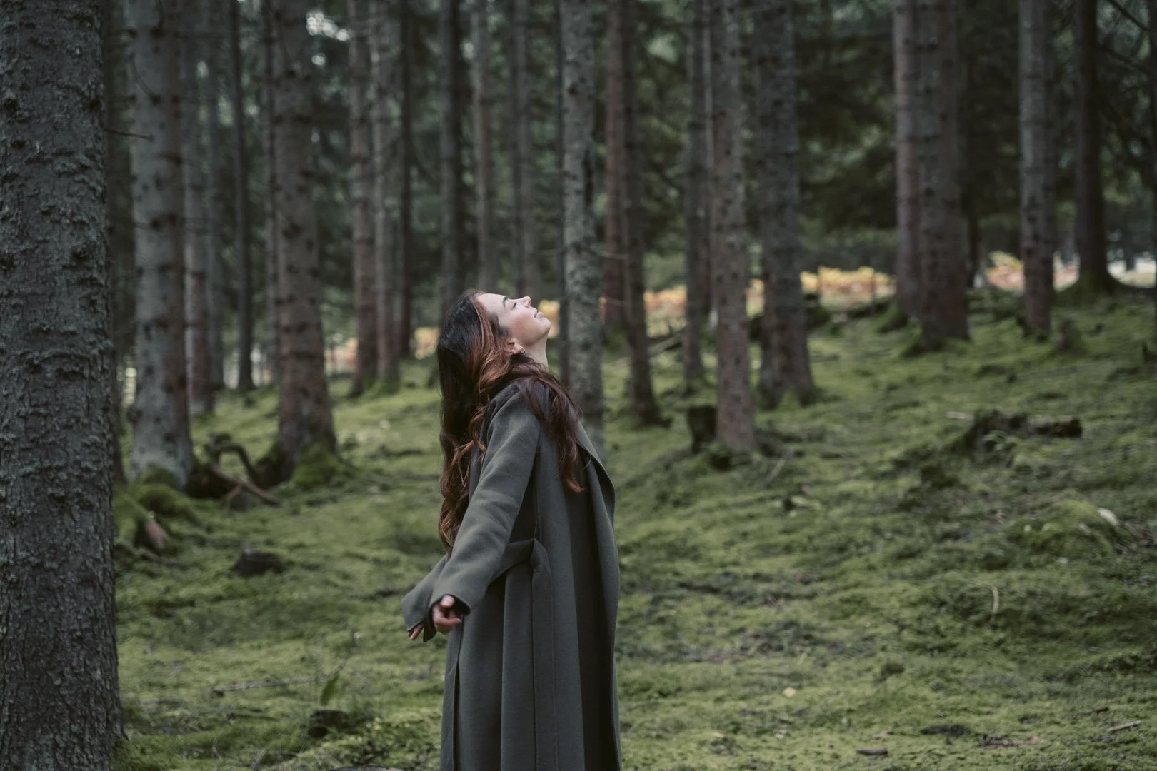 Frau in einem grünen Mantel steht in einem moosbedeckten Wald, die Augen geschlossen und das Gesicht nach oben gerichtet.